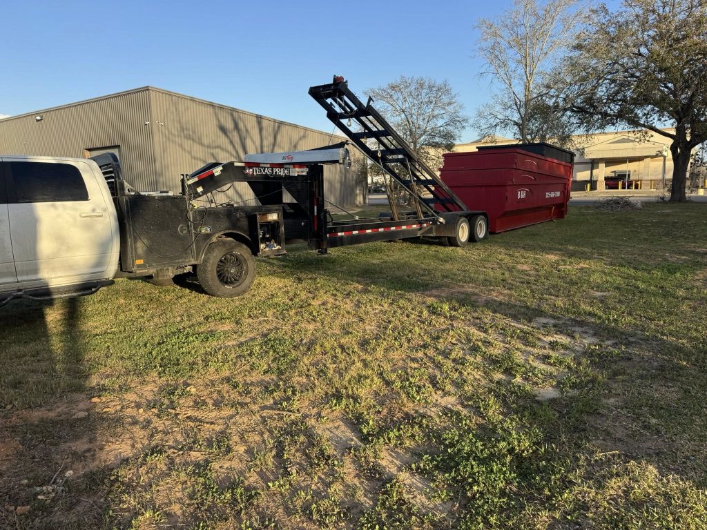 A white truck is towing a large red dumpster on a trailer in a grassy area with industrial buildings and trees in the background, under a clear sky.