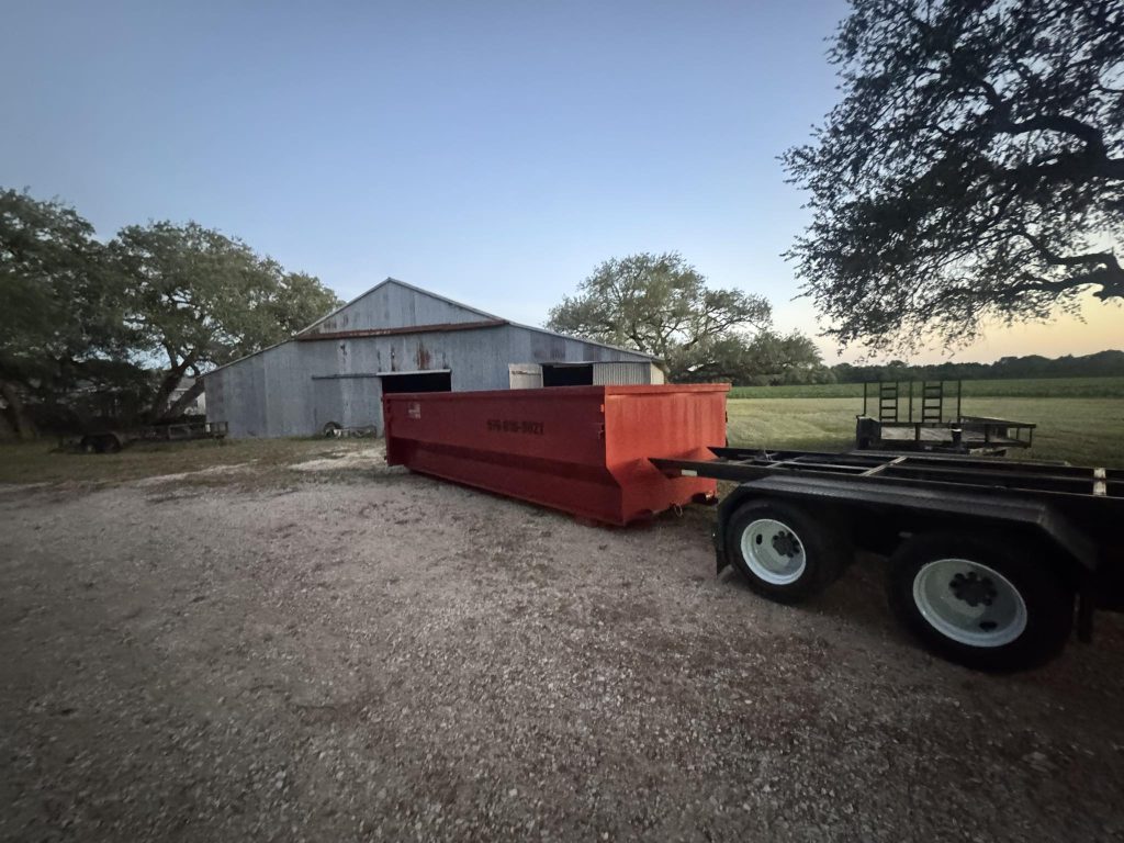 A red dumpster sits on a gravel area beside a large, rustic metal barn. Trees and open fields surround the scene under a clear evening sky.