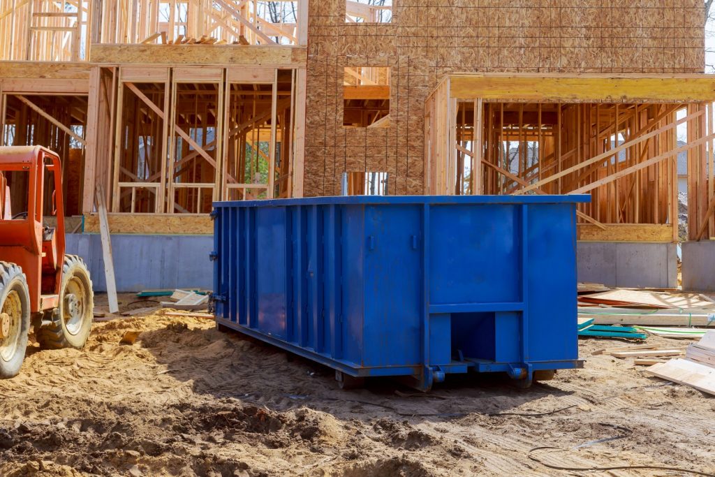A blue dumpster and red forklift are on a sandy construction site in front of a partially built wooden house, conveying a busy work atmosphere.