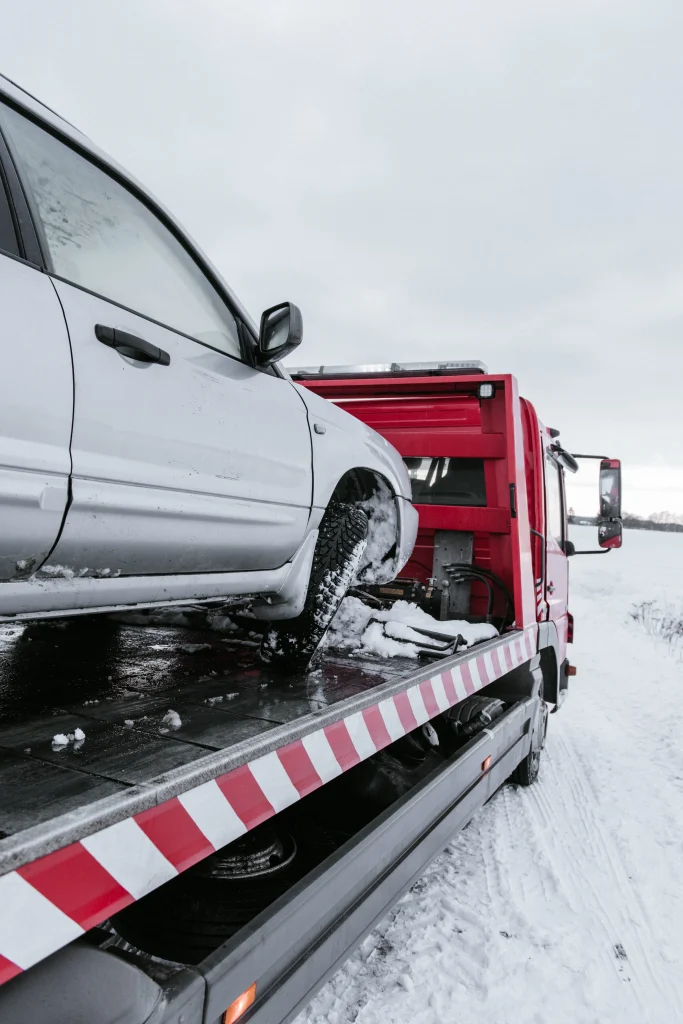 A tow truck carries a white car on its platform in a snowy landscape. The scene conveys a sense of winter emergency and road assistance.