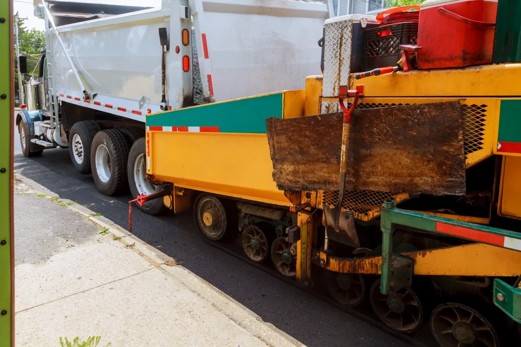 A yellow and green asphalt paver with a mounted shovel operates next to a white dump truck on a sunny street. Concrete sidewalk visible on the left.