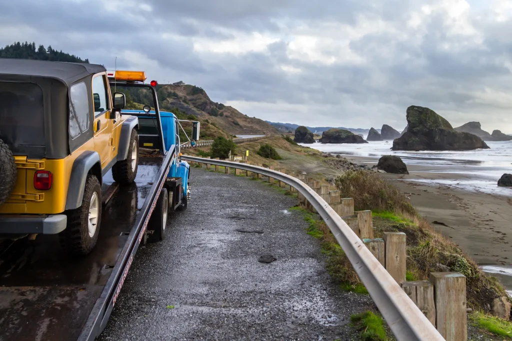 Tow truck with a yellow Jeep on a coastal road, overlooking rocky cliffs and waves. Overcast sky suggests an approaching storm. Rugged, tranquil scenery.
