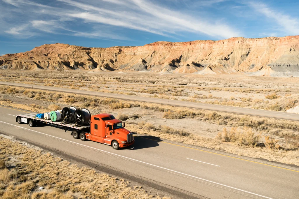 A red semi-truck with a flatbed trailer hauls large industrial reels through a vast desert landscape with rugged cliffs under a clear blue sky.