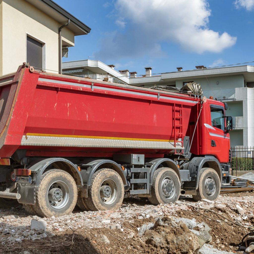 A red dump truck with multiple wheels is parked on a construction site with dirt and debris, set against a backdrop of residential buildings under a blue sky.
