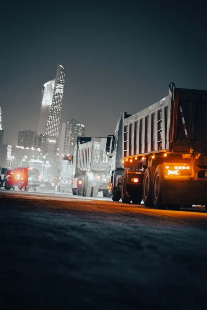 Night scene in a city with a line of trucks on a street, illuminated by streetlights. A person walks by, with tall buildings in the background.
