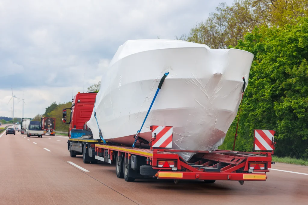 A large red truck transports a shrink-wrapped boat on a flatbed trailer down a highway. The sky is overcast, and trees line the road.